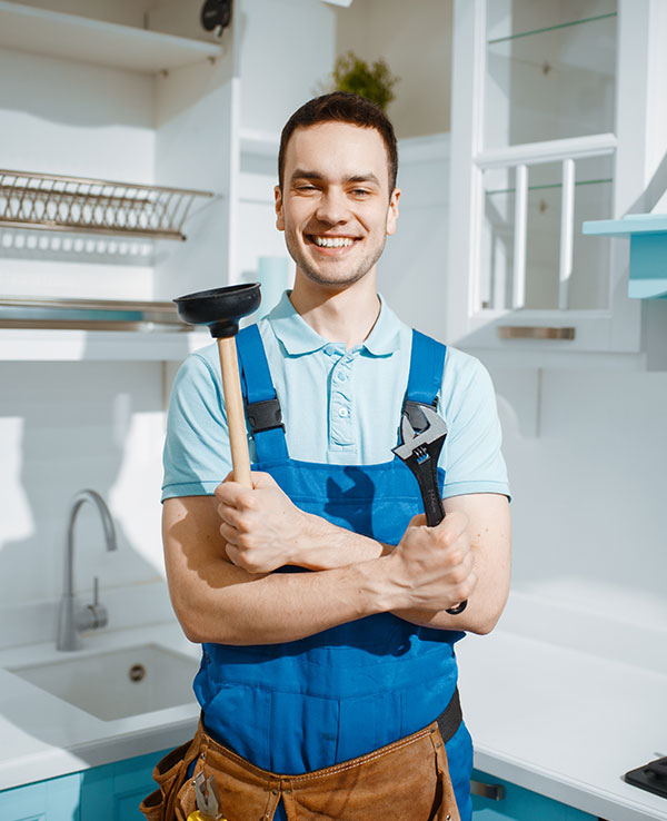 Smiling plumber in a blue uniform stands in a bright kitchen, holding a plunger and wrench. The setting conveys friendliness and reliability.