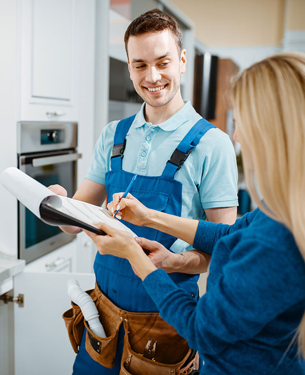 A smiling repairman in blue overalls holds a clipboard as a customer signs a document. They stand in a modern kitchen, conveying a positive, professional interaction.